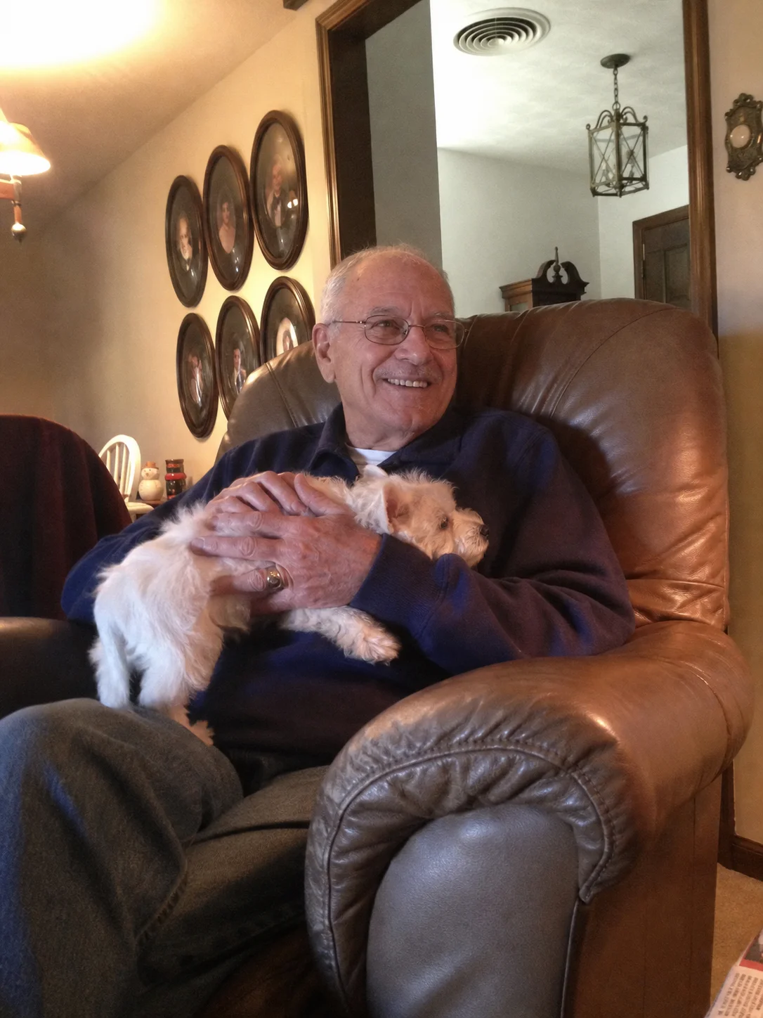 Elderly man smiling while holding his small white dog in a leather armchair